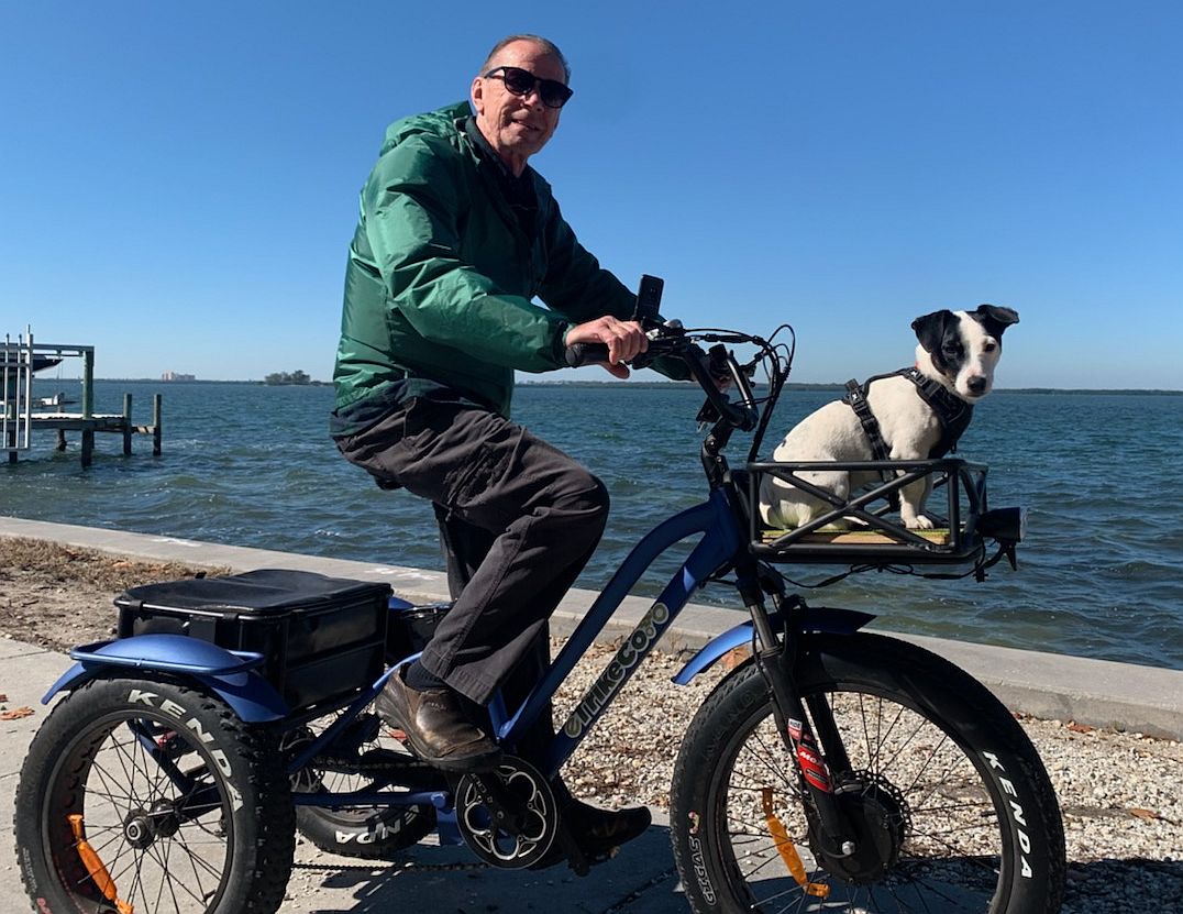 A guy sitting on a trike with his dog in the front basket in front of a body of water.